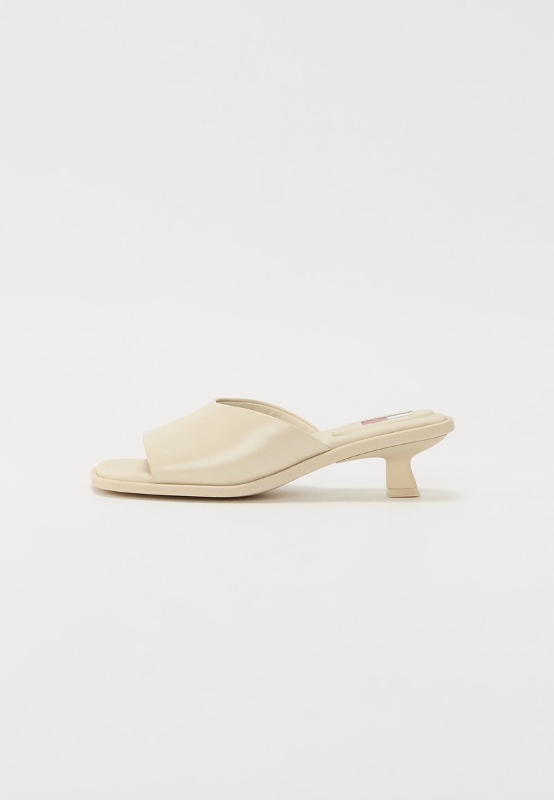 Sandale mule beige à bout ouvert avec un petit talon bloc et une large bride rembourrée sur fond blanc uni.