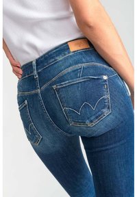 Un jean en denim bleu foncé présente une taille mid-rise, une silhouette ajustée et des coutures de poche arrière distinctes en motif de feuille. Étiquette en cuir marron à la taille.