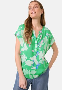 Grüne kurzärmelige Bluse mit floralem Druck in Hellrosa und Blau. Knopfkragen und glattes, leichtes Material. Abgerundeter Saum.