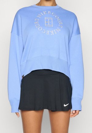 Kobieta ubrana w jasnoniebieską bluzę Nike Court i czarną tenisową spódniczkę Nike z białym logo swoosh na krawędzi spódniczki.