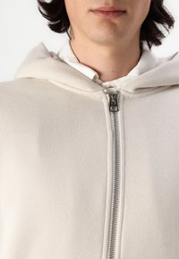 Light beige hoodie made of soft fabric, featuring a full zipper front, a drawstring hood, and a smooth texture. Simple design with no patterns.