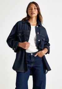 Mörk denimjacka med stora framfickor, knappstängning och uppvikta ärmar. Kombinerad med högmidjade jeans och en vit topp.