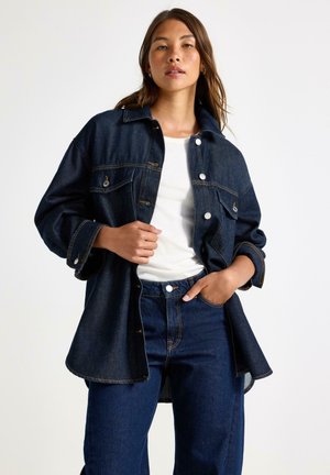 Mörk denimjacka med stora framfickor, knappstängning och uppvikta ärmar. Kombinerad med högmidjade jeans och en vit topp.