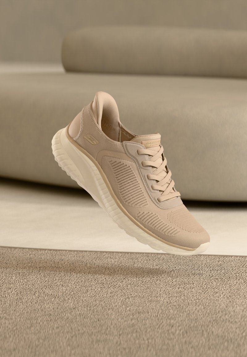 Μπεζ ελαφρύ αθλητικό παπούτσι με υφασμάτινο επάνω μέρος με υφή, παχύ μαλακό πέλμα και κορδόνια, τοποθετημένο πάνω από μπεζ χαλί και καναπέ σε απαλό φωτισμό.