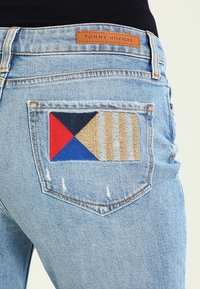 Jeans di denim azzurro chiaro con una tasca posteriore decorata da ricami multicolori in rosso, blu, nero e tonalità beige, con motivi geometrici.