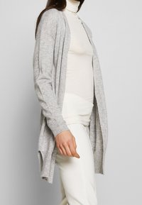 Ljusgrå kofta med öppen framkant, ribbade ärmslut och en mjuk textur, över en krämfärgad polotr öja och vita byxor.