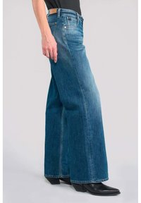 Vaqueros de denim azul de pierna ancha con un lavado desvanecido, que presentan una cintura alta, cinco bolsillos y detalles de costura. Combinados con botas negras de punta afilada.