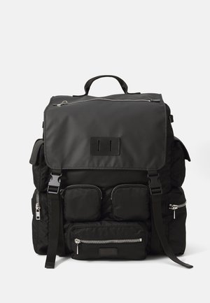 Mustmust mustkott musta nailonist, millel on klapiga sulgemine, mitu lukuga taskut, reguleeritavad rihmad ja ülemine käepide kandmisvõimaluste jaoks.