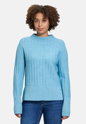 Pull en maille côtelée bleu clair avec un col montant et des manches longues. Présente un tissu texturé et un ourlet ajusté. Associé à un jean foncé.
