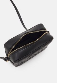 Černá prošívaná kožená kabelka přes rameno se zlatým zipem částečně otevřená, odhalující prázdný vnitřek, na bílém pozadí.