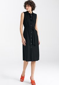 Czarna bezrękawnikowa sukienka z kołnierzykiem, zapięciem na guziki z przodu, z paskiem w talii i średniej długości krojem. Modelka ma na sobie pomarańczowe sandały na obcasie.