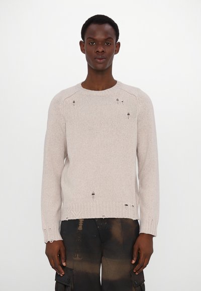 Knit sweater in light beige with a ribbed neckline and hem. Features distressed details throughout and long sleeves. Paired with dark pants.