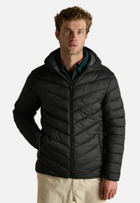 Čierna puffr jacket so kapucňou, s dizajnom kože s chevronovým prešívaním. Zips vpredu, bočné vrecká a mäkký povrch. Nosená cez zelenú košeľu.