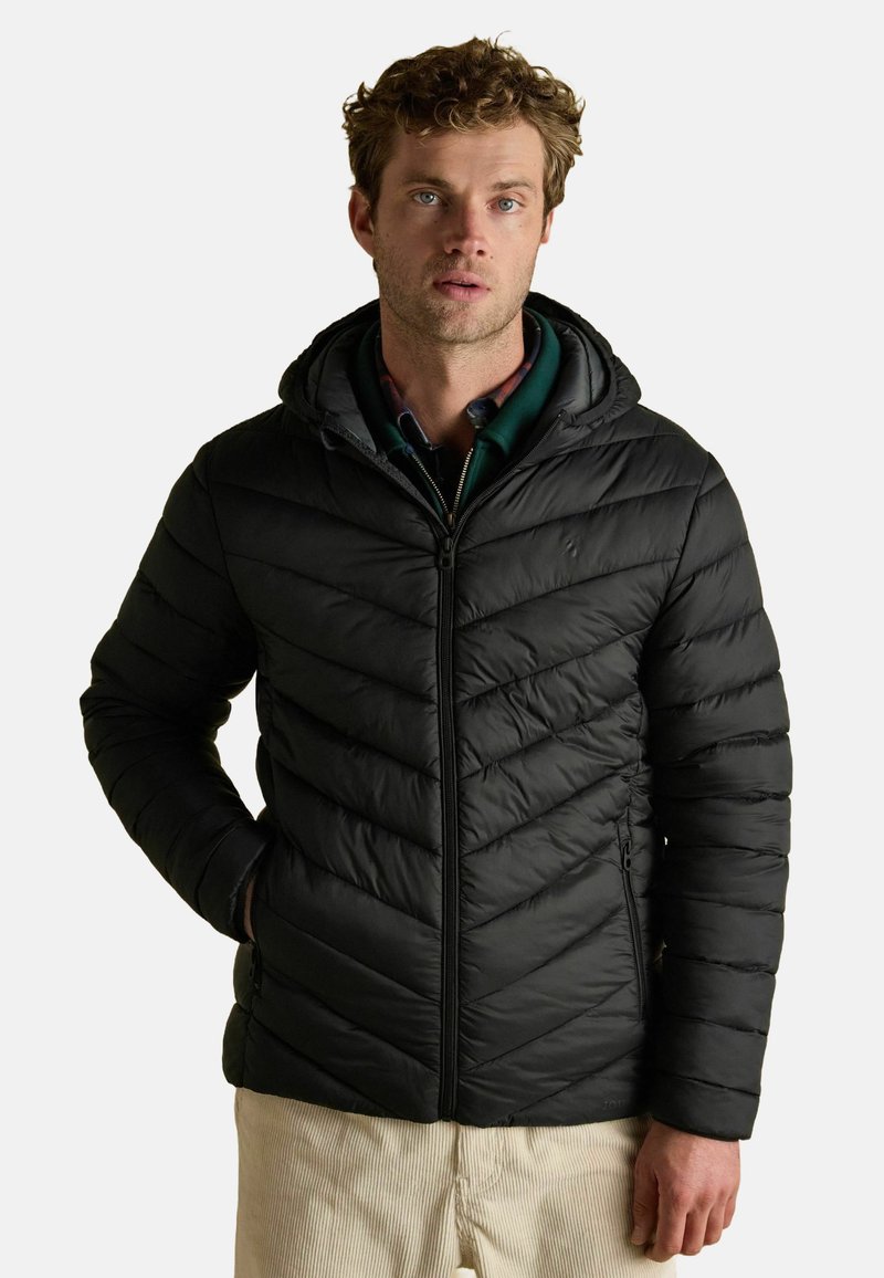 Čierna puffr jacket so kapucňou, s dizajnom kože s chevronovým prešívaním. Zips vpredu, bočné vrecká a mäkký povrch. Nosená cez zelenú košeľu.