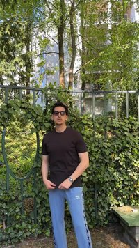Άνδρας με γυαλιά ηλίου, μαύρο t-shirt και μπλε αθλητική φόρμα Umbro στέκεται μπροστά από φράχτη καλυμμένο με κληματαριά, με δέντρα και κτίρια πίσω.