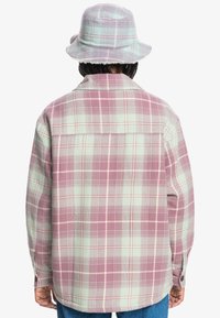 Rutigt skjorta i mjukt material, med en blandning av dämpade rosa och gröna nyanser, och en krage som knäpps ner. Kompletteras av en matchande bucket hat.