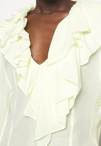 Närbild av en mörkhyad person som bär en ljusgul blus med lager av volanger runt en djup V-krage och transparenta ärmar.