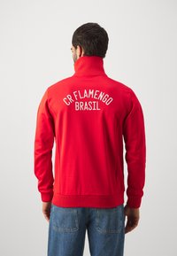 Red track jacket with high collar, ribbed cuffs and hem. Features white text: "CR FLAMENGO" and "BRASIL" on the back. Soft texture.