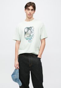 Ανοιχτοπράσινο βαμβακερό μπλουζάκι με γραφικό σχέδιο που απεικονίζει ένα σπίτι, φαντασμαγορική φιγούρα και αστέρια. Συνδυασμένο με μαύρο παντελόνι με ρίγες και μπλε καπέλο.