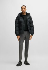 Giacca a vento nera con cuciture visibili, tasche con zip e cappuccio, abbinata a pantaloni grigi su misura e stivali alla caviglia neri.