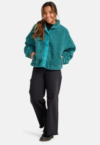 Teal fleece jack met een knoopsluiting aan de voorkant en een kraag, gecombineerd met zwarte flare-broeken met rafelige zoom en zwarte veterschoenen.