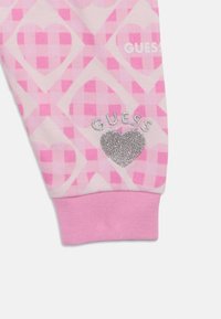 Roze stof met patroon van harten en ruiten, met een zilveren glitter "GUESS" logo en een hartaccent op de manchetten.