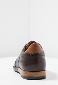 Chaussure en cuir marron avec une texture lisse, un talon beige contrastant et un bout arrondi. La semelle est en bois avec des rainures horizontales.