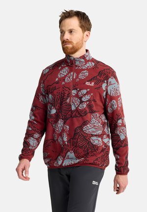 Man draagt een rood-grijs patroon fleecejack met halve ritssluiting en zwarte broek, loopt met een neutrale uitdrukking tegen een effen achtergrond.