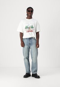 White graphic t-shirt with green and red print, light blue denim jeans, and black chunky shoes. Person wearing round sunglasses.