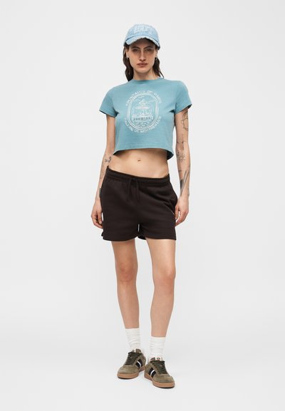 Γυναίκα που φοράει ανοιχτό μπλε κοντό μπλουζάκι με γραφικό, μαύρα σορτς, μπλε καπέλο, λευκές κάλτσες και πράσινα αθλητικά παπούτσια, στέκεται μπροστά σε ένα απλό φόντο.