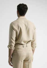 Camisa de lino beige con mangas enrolladas, combinada con pantalones beige a juego. Diseño simple con una textura sutil y sin patrones visibles.