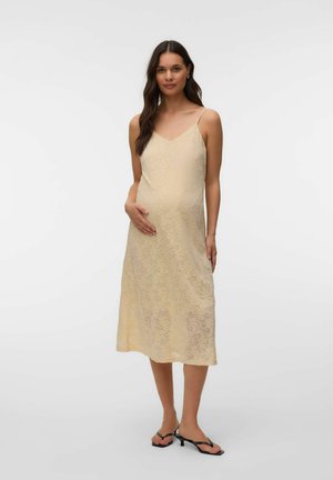 Femme enceinte aux longs cheveux foncés portant une robe nuisette midi en dentelle beige et des sandales à talons noires, debout devant un fond uni.