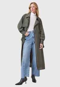 Trench coat de color verde oliva con mangas anchas y cintura ceñida con cinturón, combinado con jeans de tiro alto de color azul claro y botines negros de punta.