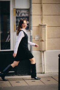 Jeune femme marchant sur un trottoir en ville, portant une robe noire par-dessus une chemise blanche et des bottes noires, avec une affiche portant l'inscription "Yung Lean" en arrière-plan.