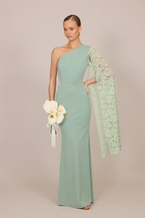 Kvinde i gulvlange mintgrøn enkeltskulderkjole med blonderm, der holder en buket af hvide calla-liljer mod en neutral baggrund.