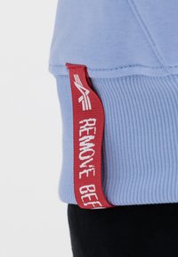 Sudadera azul claro con puños acanalados y una etiqueta roja prominente con texto blanco que dice "REMOVE BEF." Textura suave, diseño casual.