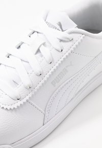 Bílé kožené tenisky s plochými bílými tkaničkami, se stříbrným logem Puma a jemnými švy podél strany a paty.