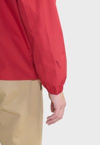 Rode lichtgewicht jas met elastische boorden, zachte textuur en een logodetail op de mouw. Gepaard met beige broeken.