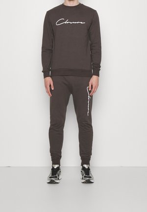 Homme portant un sweat-shirt à manches longues marron foncé et un jogging assorti avec un texte en écriture blanche, associé à des baskets argentées et noires.
