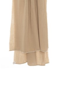 Beżowy szal z muślinu o lekkiej, pomarszczonej fakturze. Posiada surową krawędź z delikatnym przeszyciem i małą metką marki przymocowaną do tkaniny.