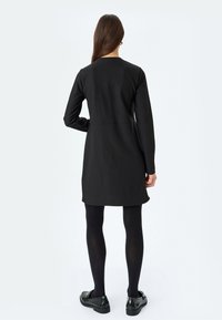 Vestito nero a maniche lunghe con silhouette dritta, taglio al ginocchio e una texture liscia. Indossato con collant neri e scarpe.