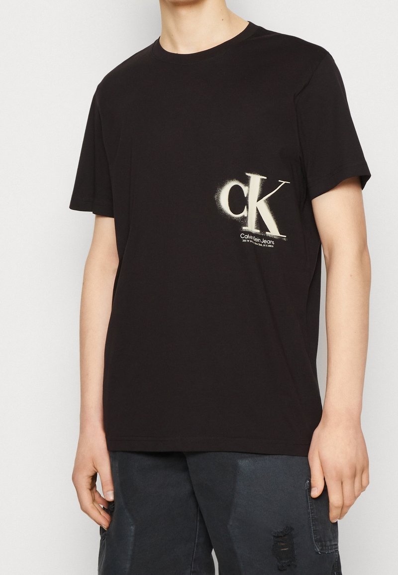 Černé bavlněné tričko s krátkým rukávem. Na přední straně se nachází velké bílé logo "CK" a text "Calvin Klein Jeans". Jednoduchý design.