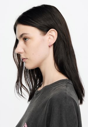 Pendientes de aro plateados exhibidos en una modelo con cabello largo y oscuro. La modelo lleva una camiseta gris oscuro. La vista en primer plano destaca el diseño de los pendientes.