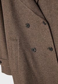 Ruskea houndstooth-kuvioinen blazer, joka on valmistettu teksturoidusta kankaasta. Takissa on kaksirivinen muotoilu, jossa on neljä mustaa nappia ja sivutasku.