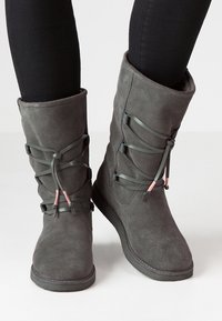Bottines en suède gris à tige haute, avec des lacets décoratifs et des accents roses. Texture lisse et semelle plate en caoutchouc.