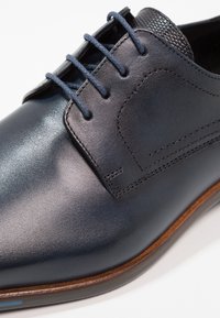 Dark blue leather shoe with smooth texture, featuring flat laces, subtle stitching, and a brown leather sole. Accented with a textured panel.