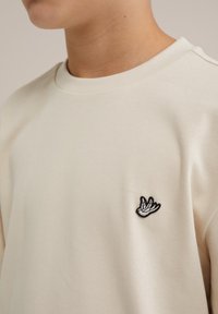 Beige pulóver kerek nyakkal, bal mellrészen egy texturált fekete-fehér hímzett kézmotívummal.