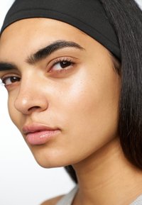 Nærbilde av et ansikt med jevn, lys brun hud og fyldige lepper. Håret er langt og rett, delvis dekket av et svart pannebånd.