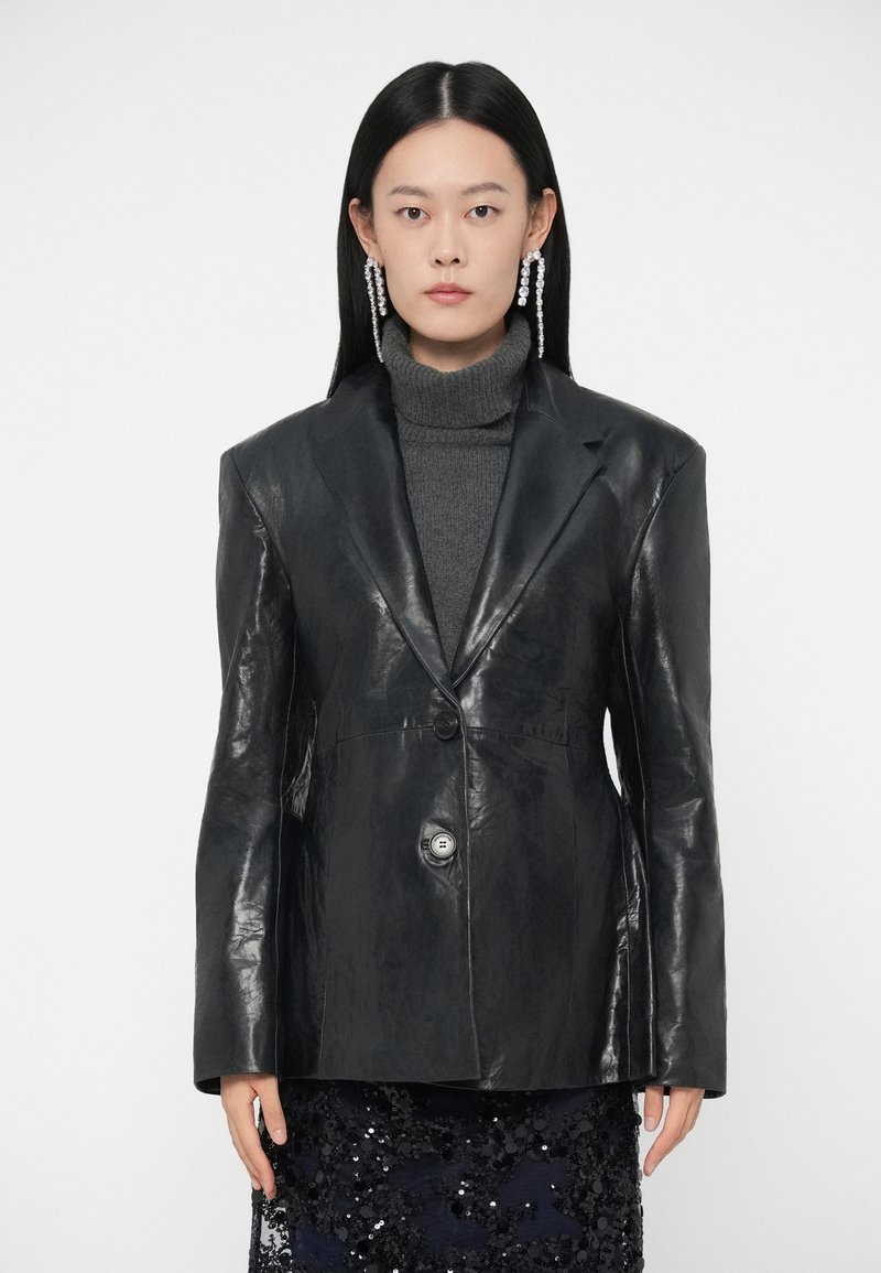 Czarna skórzana marynarka o wyraźnej sylwetce, zapinana na jeden guzik, z ostrymi klapami, nałożona na szary sweter z golfem.