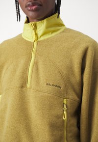 Close-up of person wearing a mustard yellow Salomon fleece pullover with a front zipper and zippered pocket.